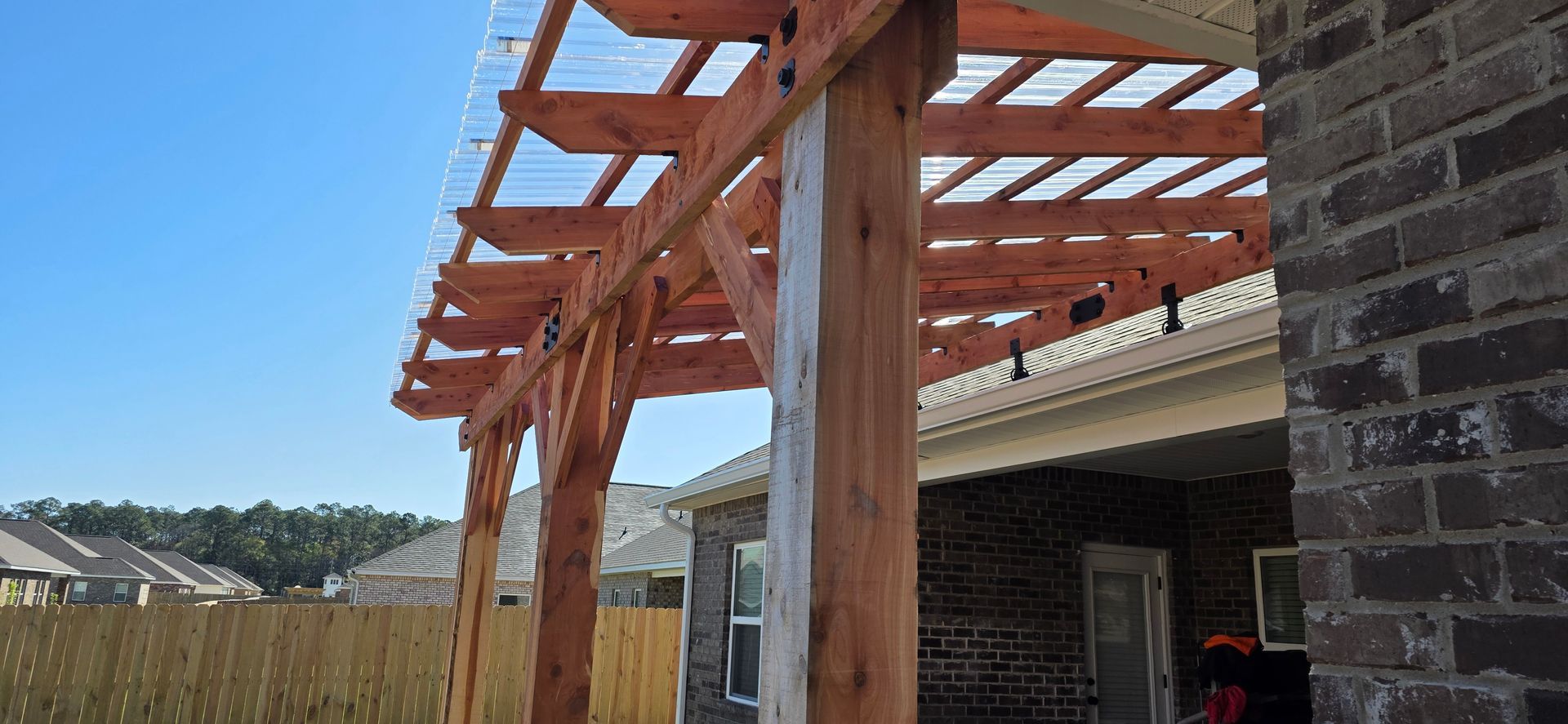 A wood pergola with a translucent roof attached to the brick wall of a house under a clear blue sky.