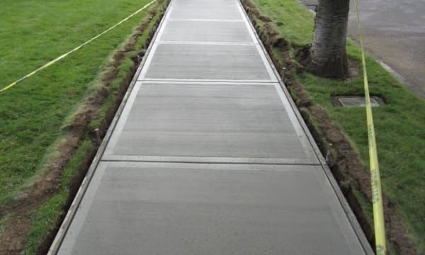 A freshly poured, light gray concrete sidewalk stretches into the distance, bordered by green grass and caution tape.