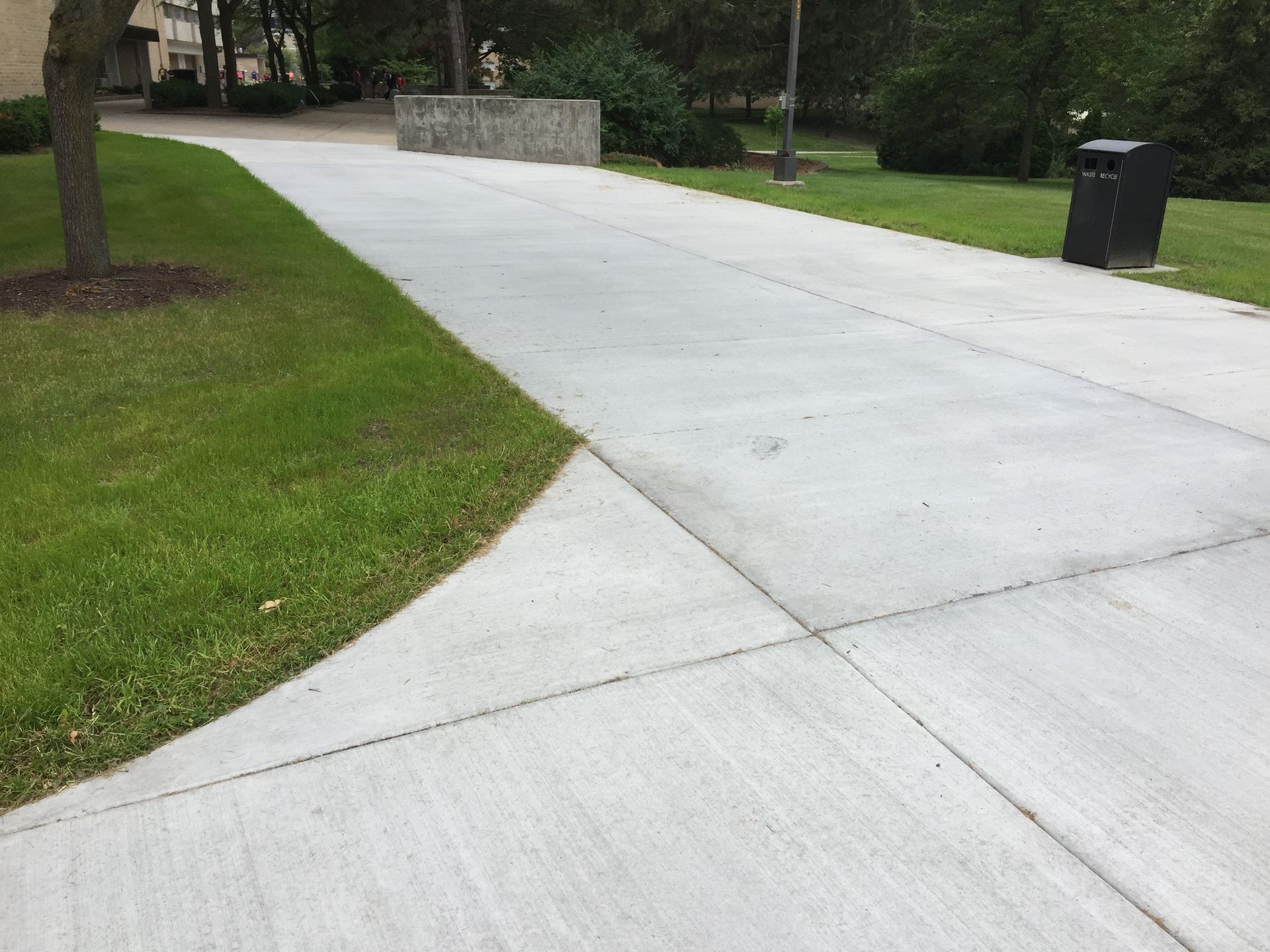 A wide, newly paved concrete walkway curves through a grassy park area near a stone wall and a trash receptacle.