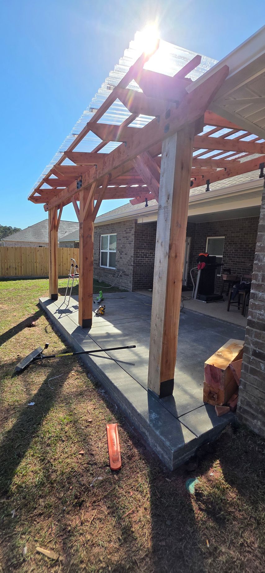 A wooden pergola with a clear corrugated roof attached to a brick house over a dark patio, viewed on a sunny day.