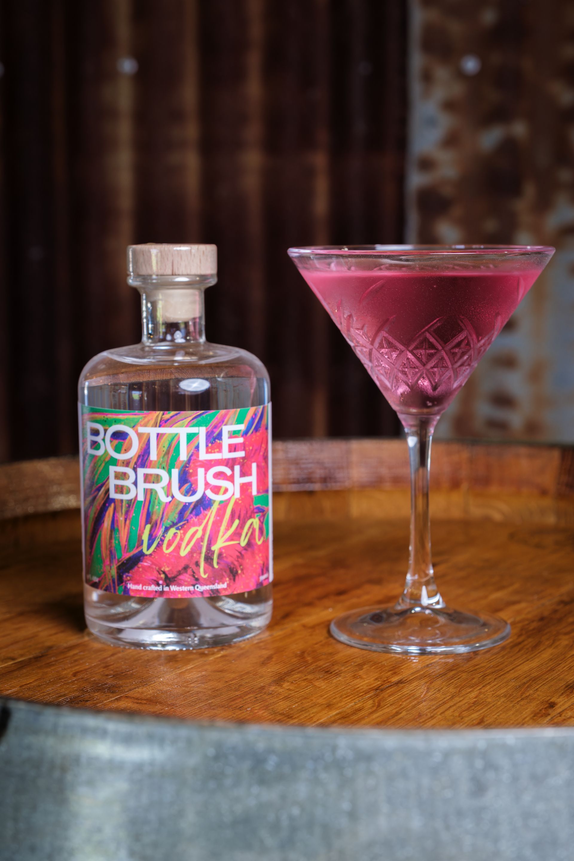 A bottle of Bottlebrush Vodka beside a pink cocktail served in a martini glass on a wine barrel.