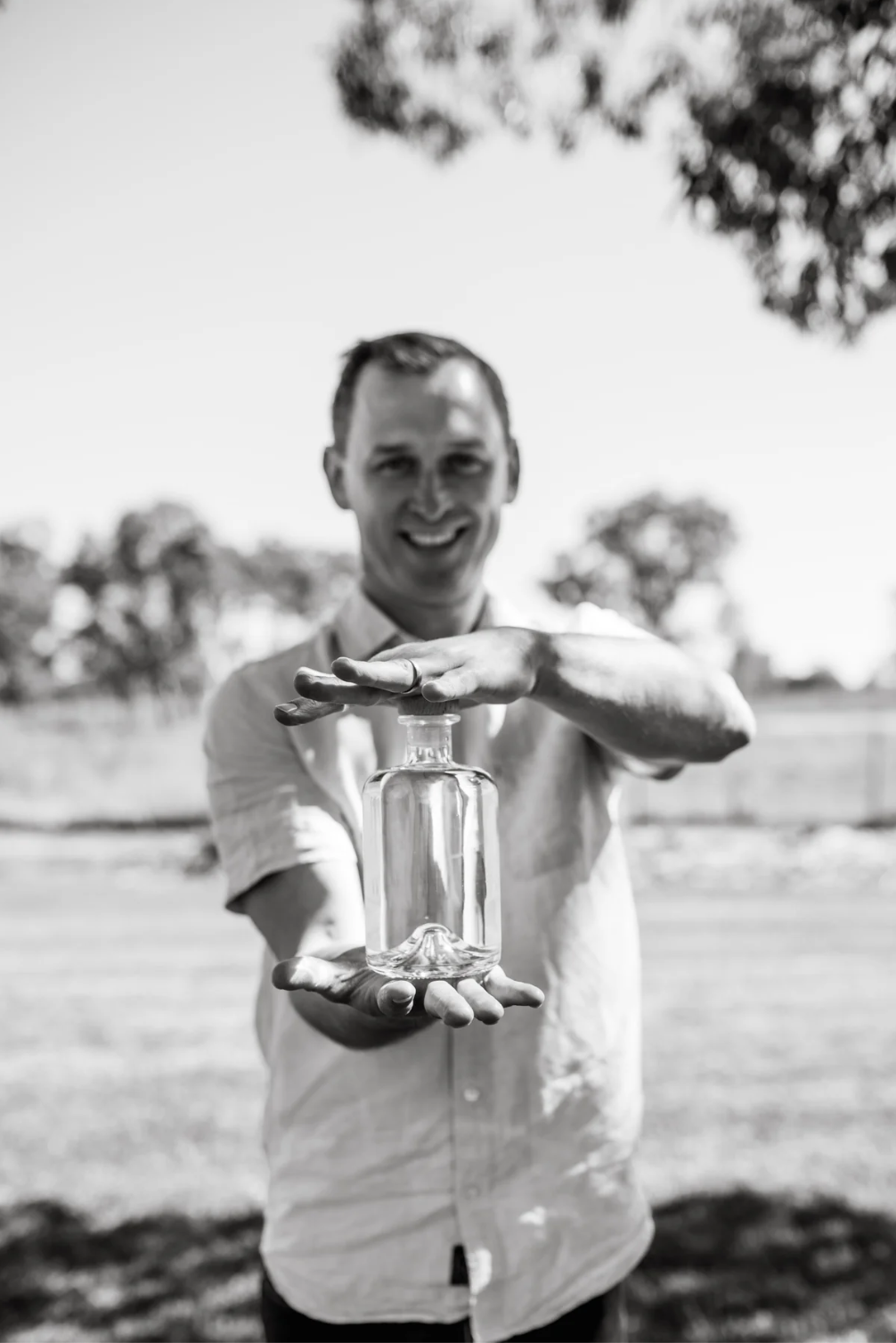 Will Fellowes, the owner of Western Queensland Spirit stands in a field holding a bottle of gin. 