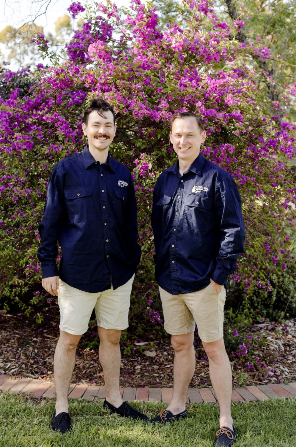 Two men are standing next to each other in front of a bush with purple flowers.