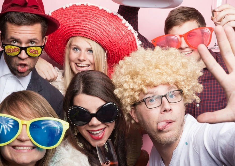 Six people posing playfully in colorful party hats and oversized glasses, smiling at the camera.