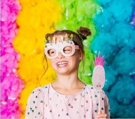 Person wearing a unicorn mask smiles in front of a bright rainbow cloud backdrop.