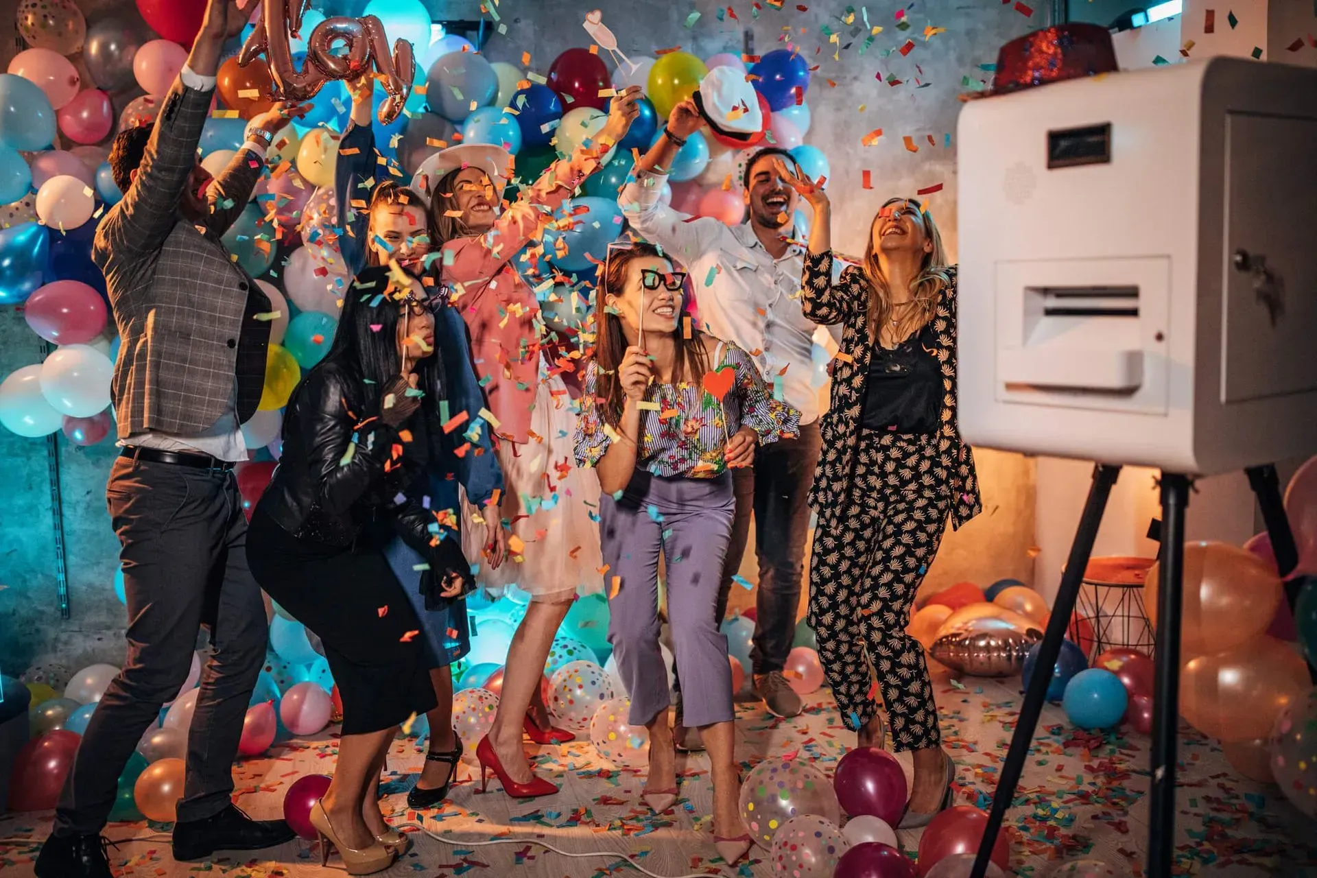 People dancing and cheering at a colorful confetti-filled party beside a photo booth