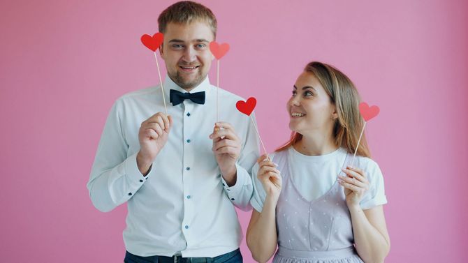 Man in white shirt and woman in pink dress hold heart props against a pink background, smiling at each other