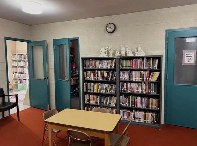 Quiet library corner with bookshelves, table and chairs, teal doors, and a wall clock.