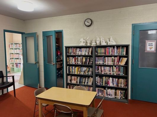 Quiet library corner with bookshelves, table and chairs, teal doors, and a wall clock.