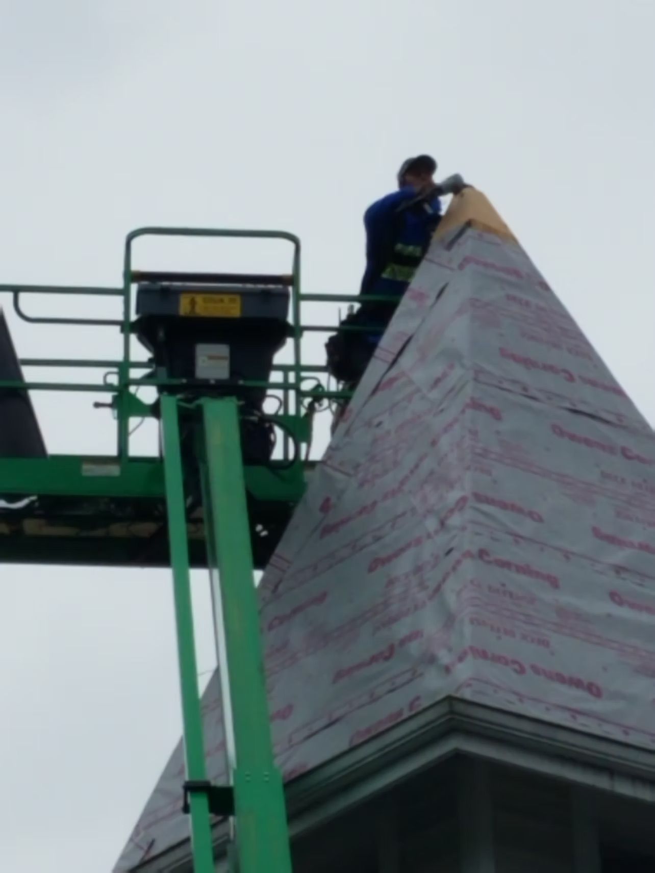 Worker on a green lift installing wrap on a tall pyramid-shaped structure