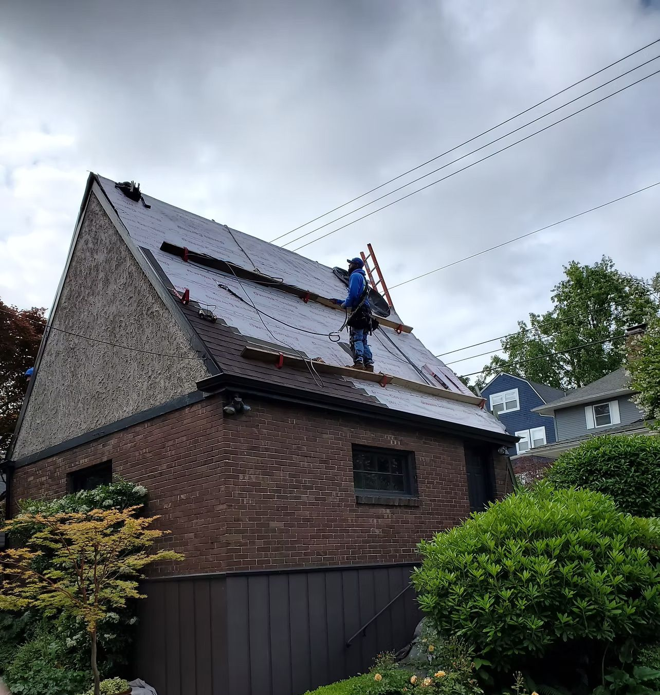 Worker on a steep roof installing shingles on a brick house under a cloudy sky