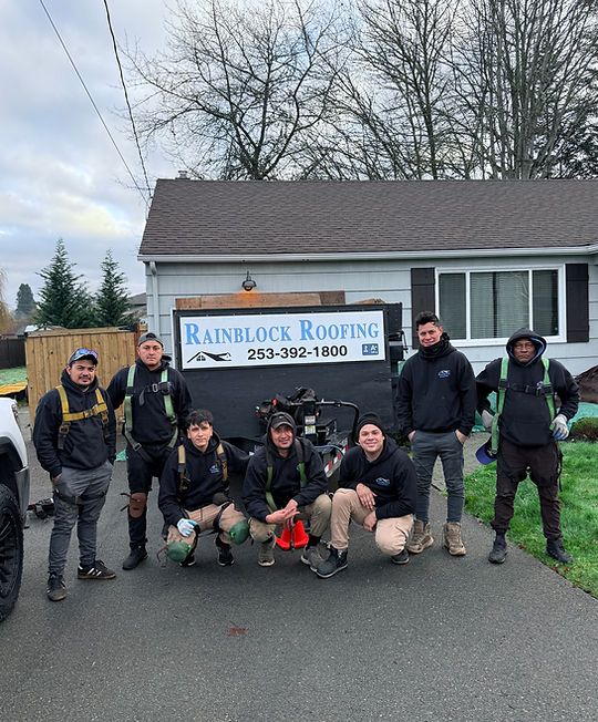 Seven people pose outside a house beside a Rainbow Roofing truck on a rainy day.