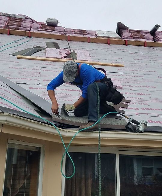 Worker kneels on a roof, fixing shingles near the edge with tools and a green cord nearby.