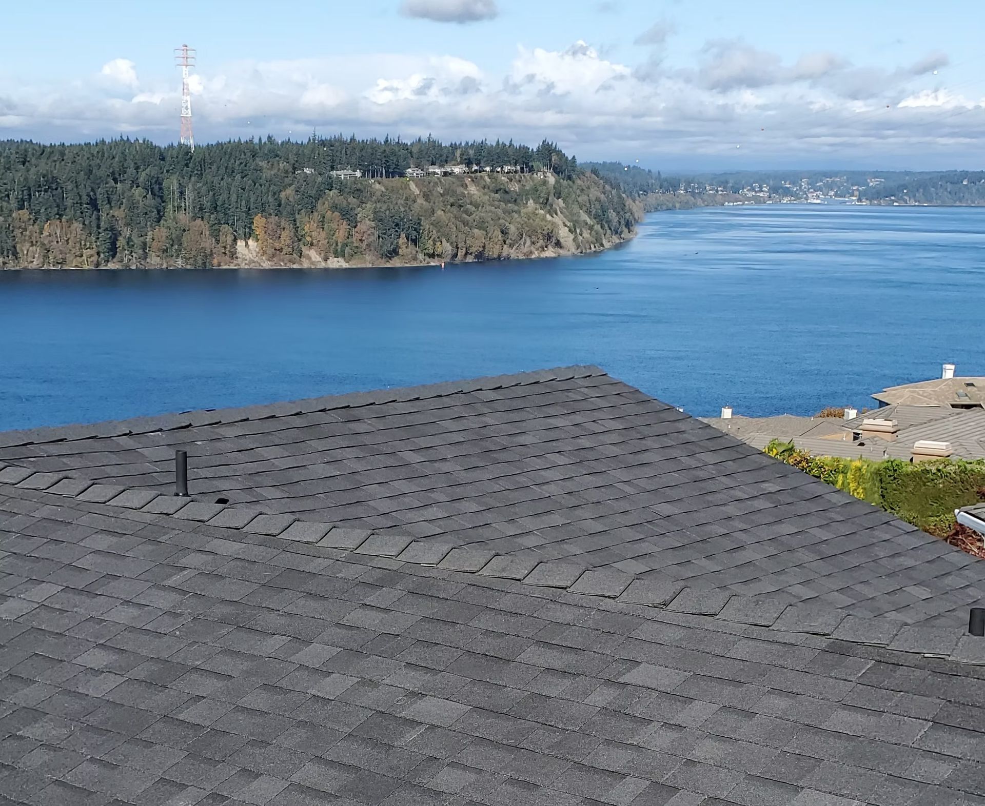 Dark gray rooftop overlooking a blue bay, tree-covered shoreline, and distant city under a partly cloudy sky
