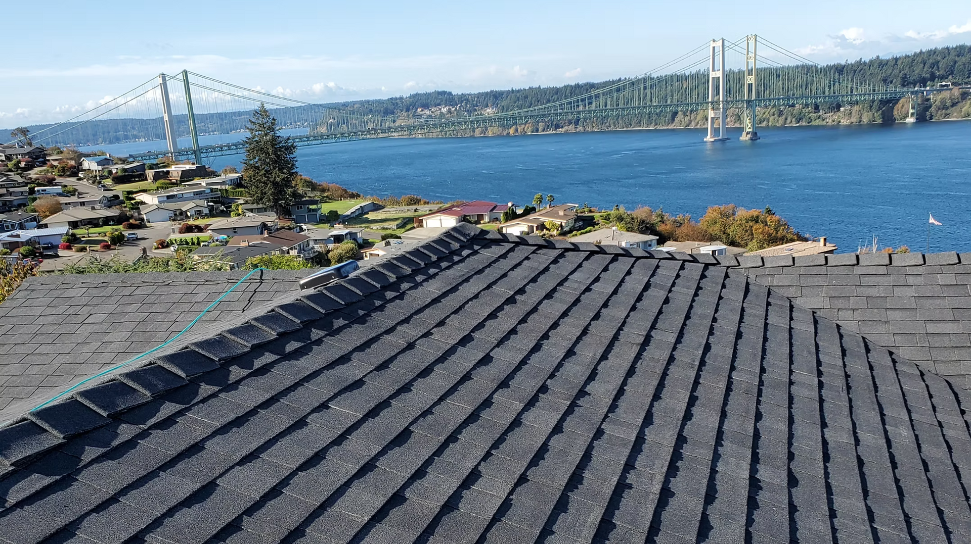 Roof shingles in the foreground overlooking blue water, a distant bridge, and a city skyline under clear skies