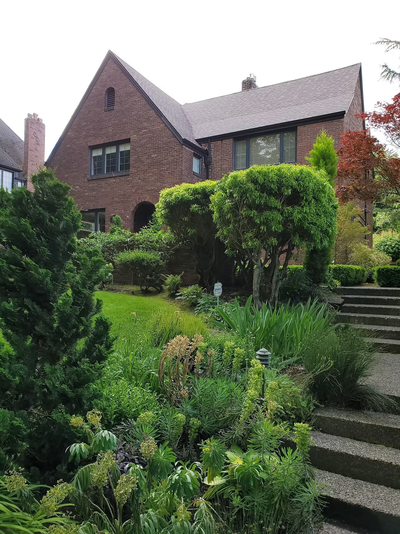 Brick house with a steep roof, lush front garden, and stone steps leading up the lawn.