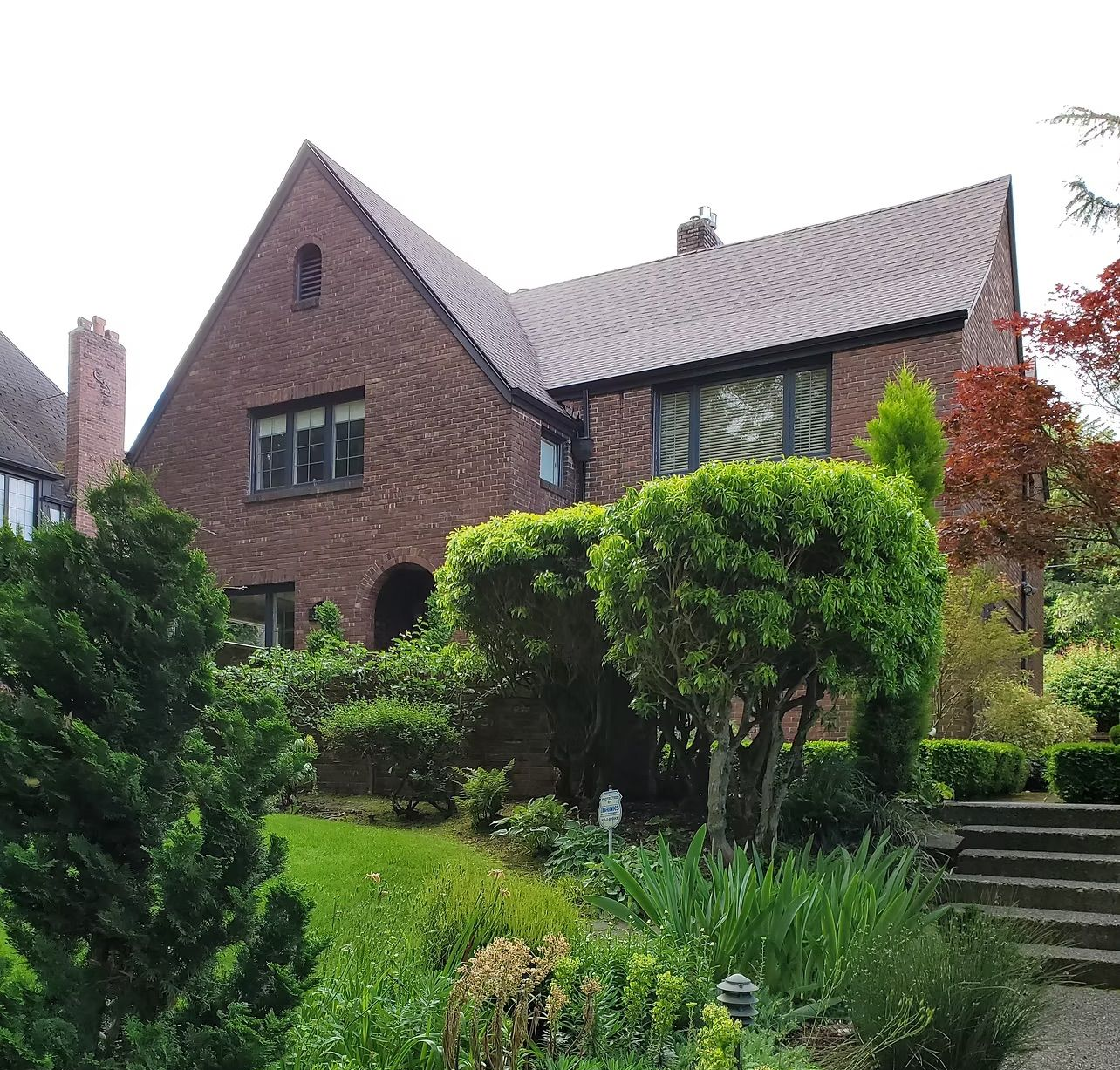 Brick house with a steep roof, surrounded by lush green shrubs and a garden.