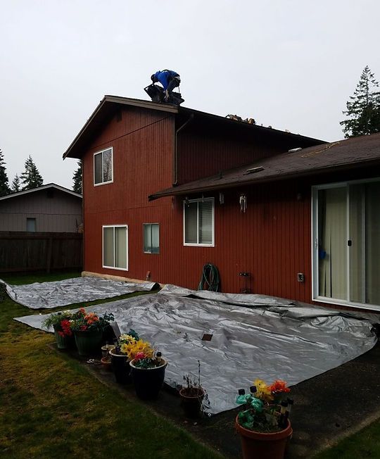 Worker on a roof above a red house, covering the yard with plastic sheeting.