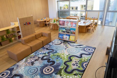 Bright daycare classroom with colorful rug, small tables, chairs, and toys by large windows
