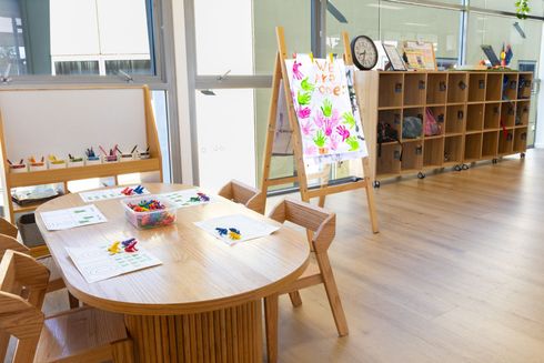 Childcare classroom with round table, art supplies, easel, and cubby shelves by a window