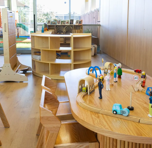 Wooden toy play area with a circular activity table, small vehicles, and colorful figurines in a bright room