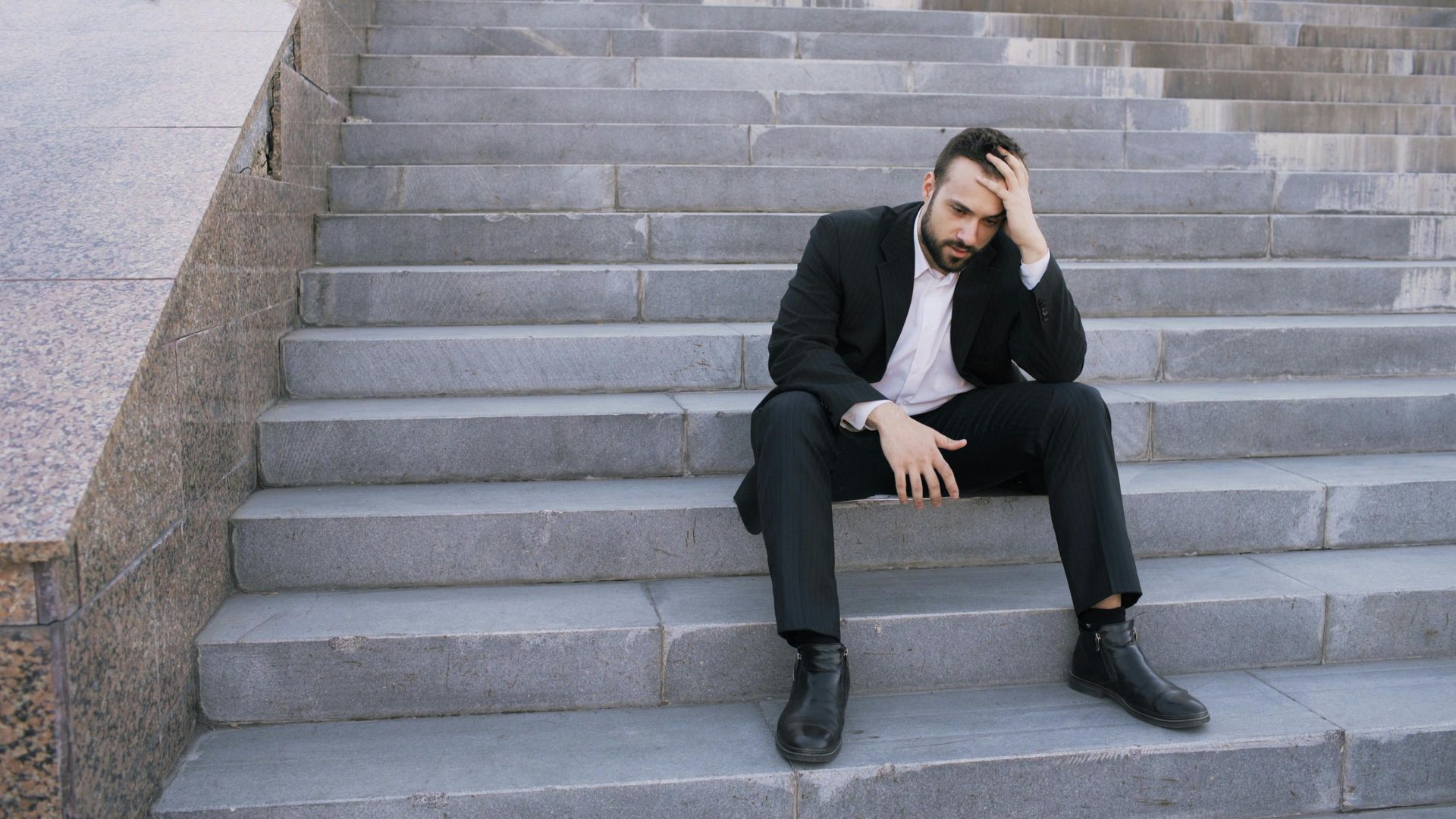 Man in black suit sitting on stone steps, holding his head with a stressed expression.