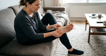 Woman sitting on a gray sofa, rubbing her foot in a living room, suggesting foot pain or discomfort