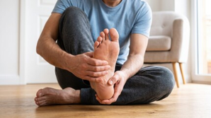 Person sitting on a hardwood floor, holding the sole of one foot toward the camera in a living room.