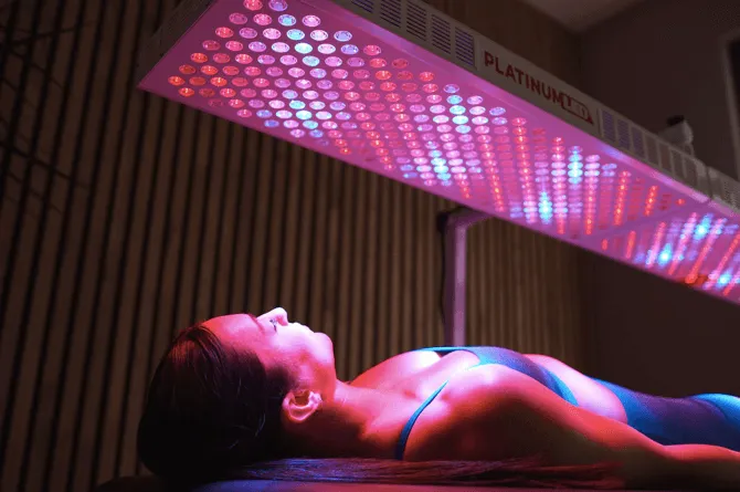 Person lying under a pink-and-blue LED tanning bed in a dim room