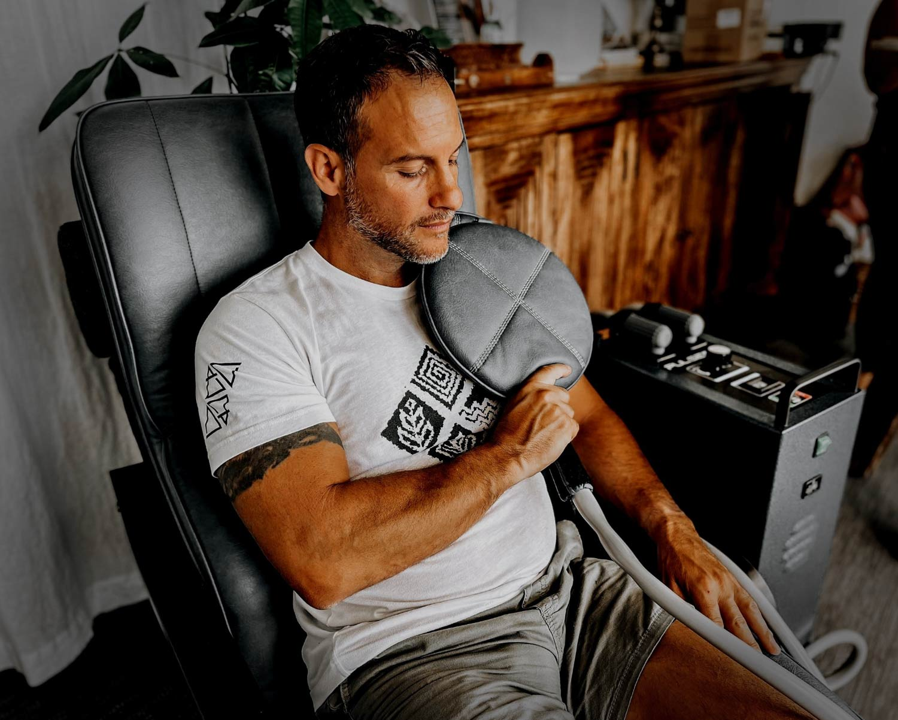 Person using a gray massage gun on their upper arm while sitting in a chair indoors