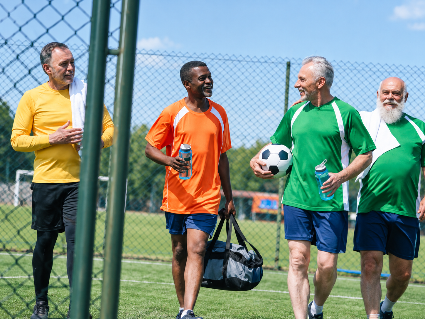 Four men in sportswear walking on a soccer field, carrying a ball and gear by a chain-link fence.