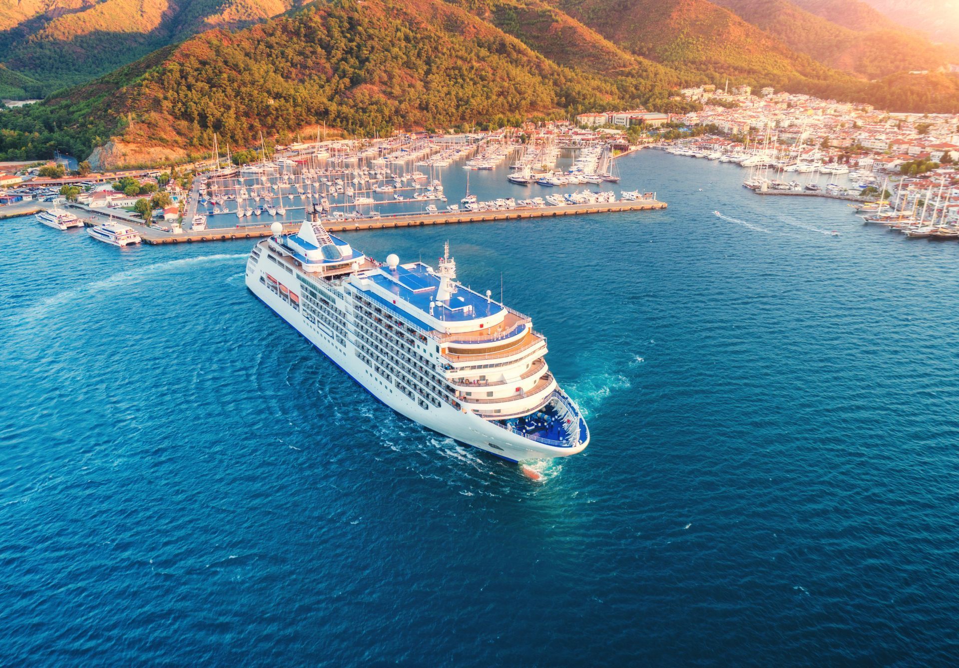 Cruise ship sailing out of a harbor, with mountains in the background.