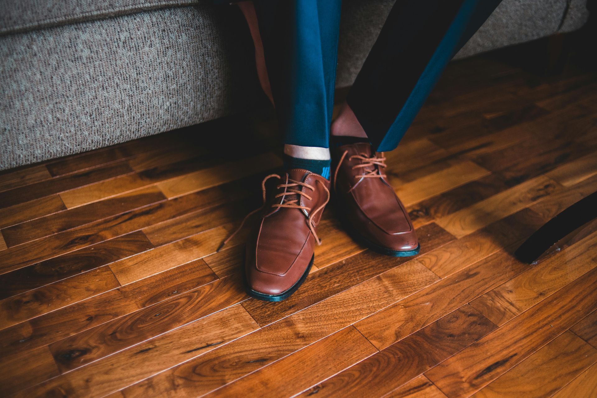 Scarpe di pelle marrone su un pavimento di legno, pantaloni blu in vista, accanto a un divano grigio.
