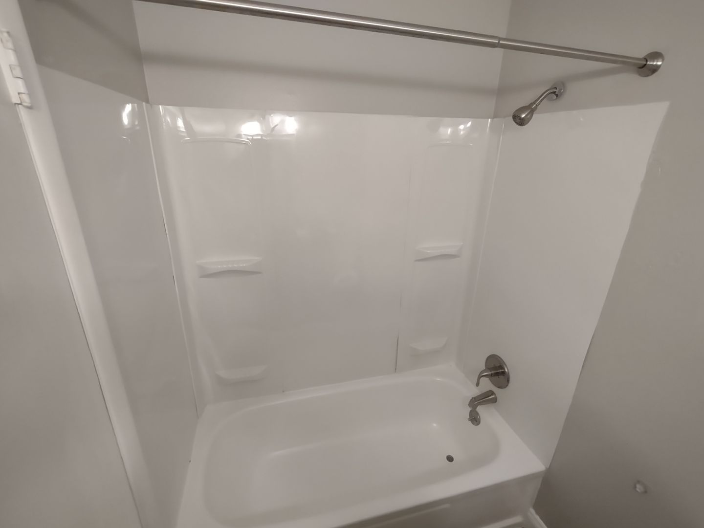 A white bathtub and shower surround with chrome fixtures and a shower curtain rod in a neutral-toned bathroom.