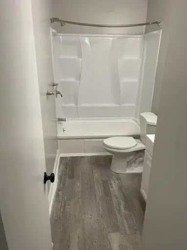 A modern bathroom featuring grey wood-look flooring, a white toilet, and a white bathtub with a shower surround.
