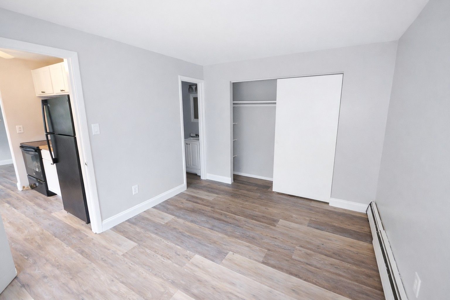 A light gray room with wood-look flooring, showing a kitchen doorway, a hallway, and a closet with sliding white doors.