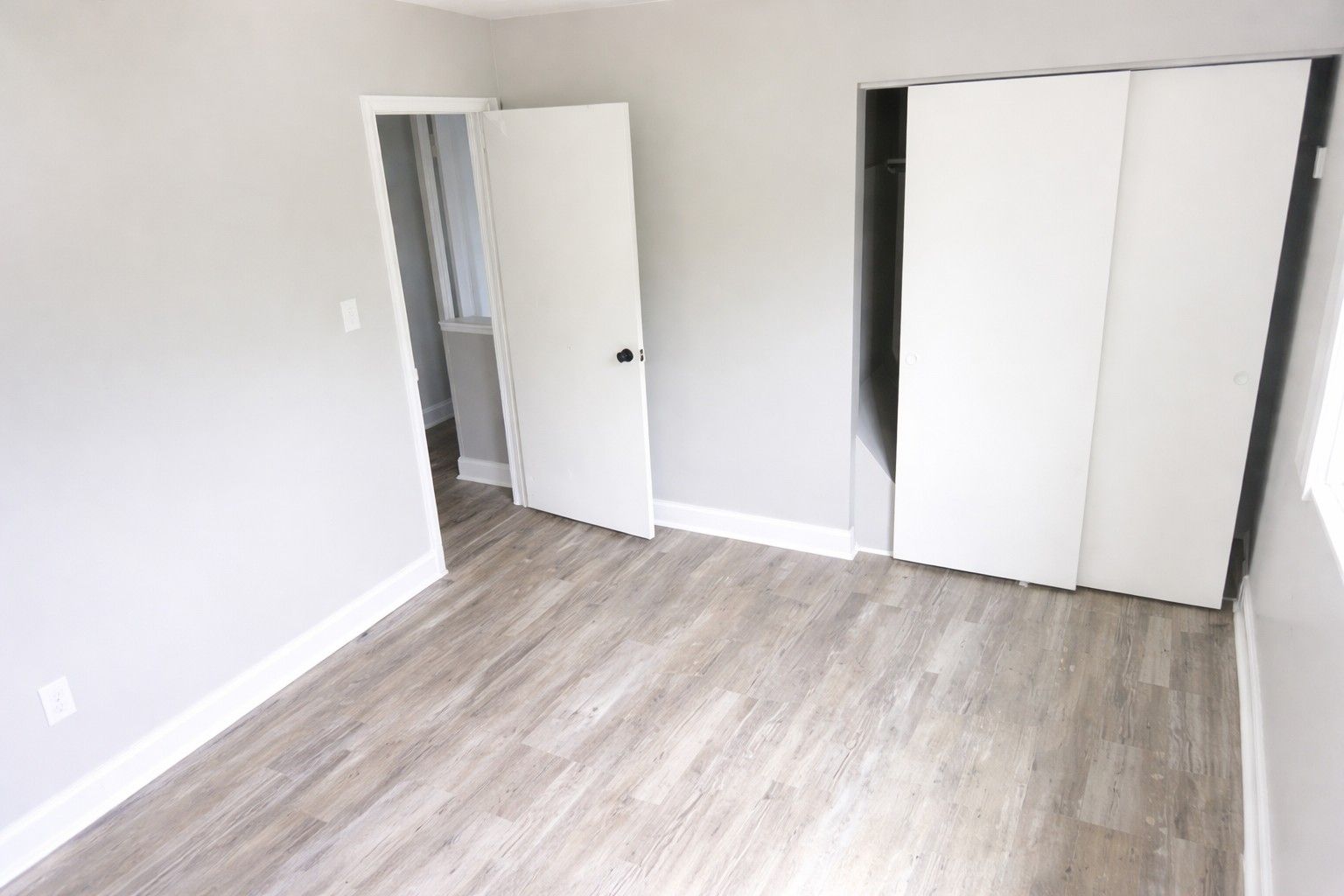 An empty bedroom with light gray walls, wood-look flooring, a white interior door, and a white sliding-door closet.