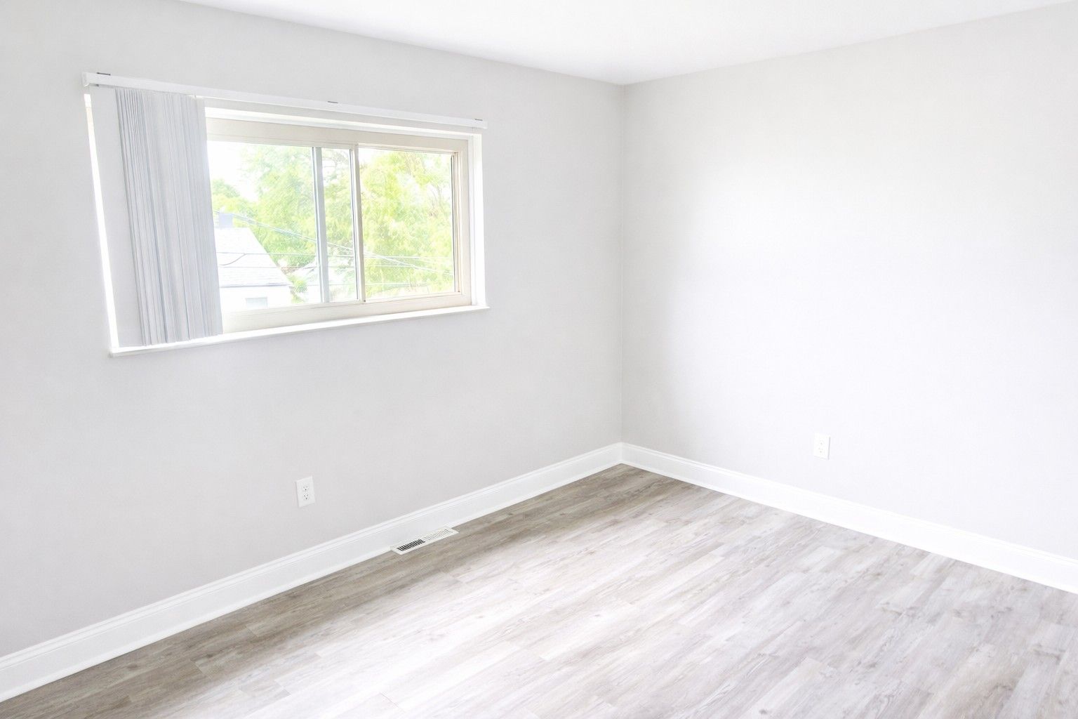 An empty room with white walls, a single window with blinds, and light-colored wood-look flooring.