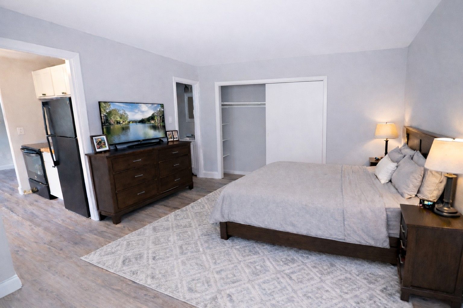 A furnished bedroom with gray walls, a bed, a dresser with a TV, a rug, and an adjacent kitchenette.