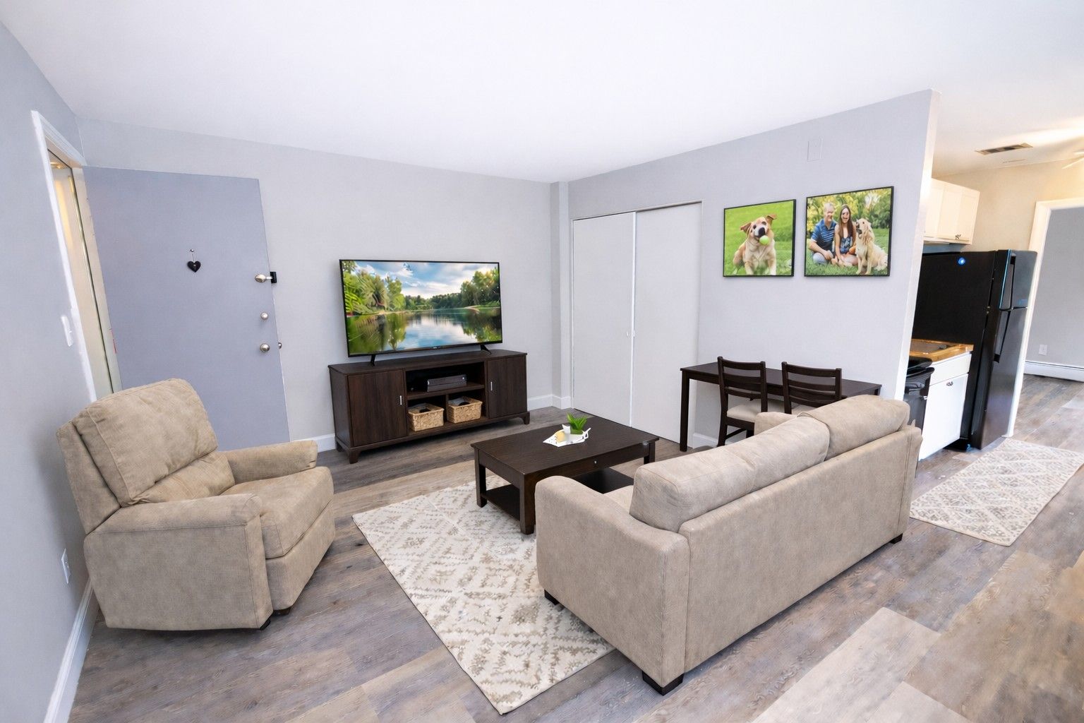 Living room with a tan armchair, sofa on a patterned rug, media console, TV, and small dining area near the kitchen.