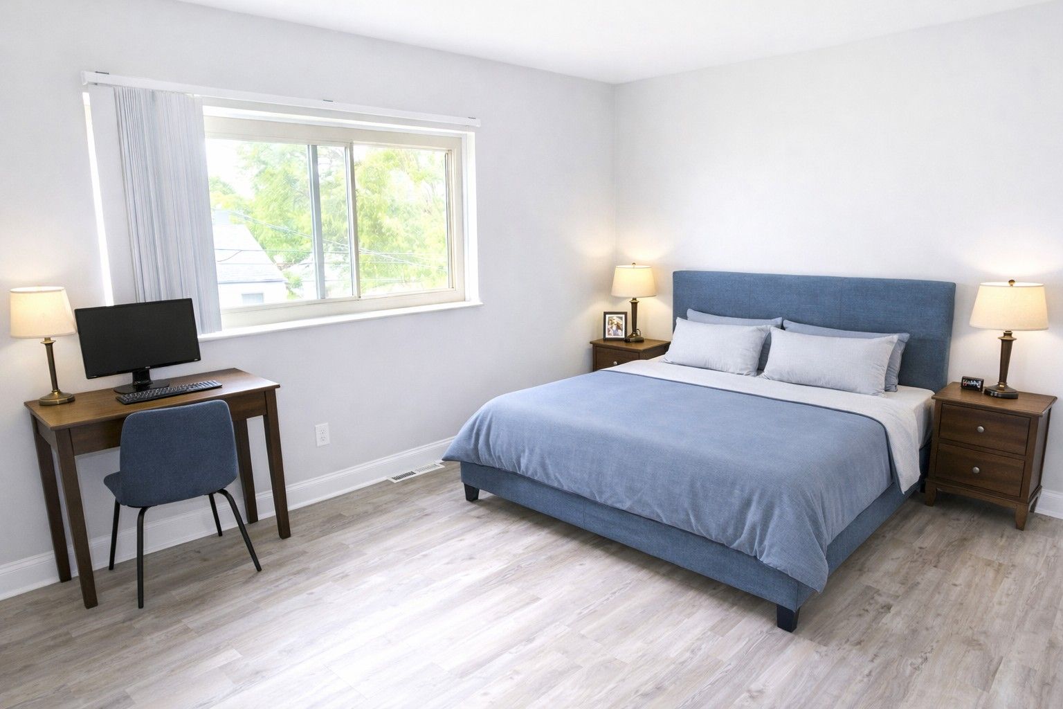 A bright, modern bedroom featuring a blue upholstered bed, a matching desk with a computer, and two wooden nightstands.