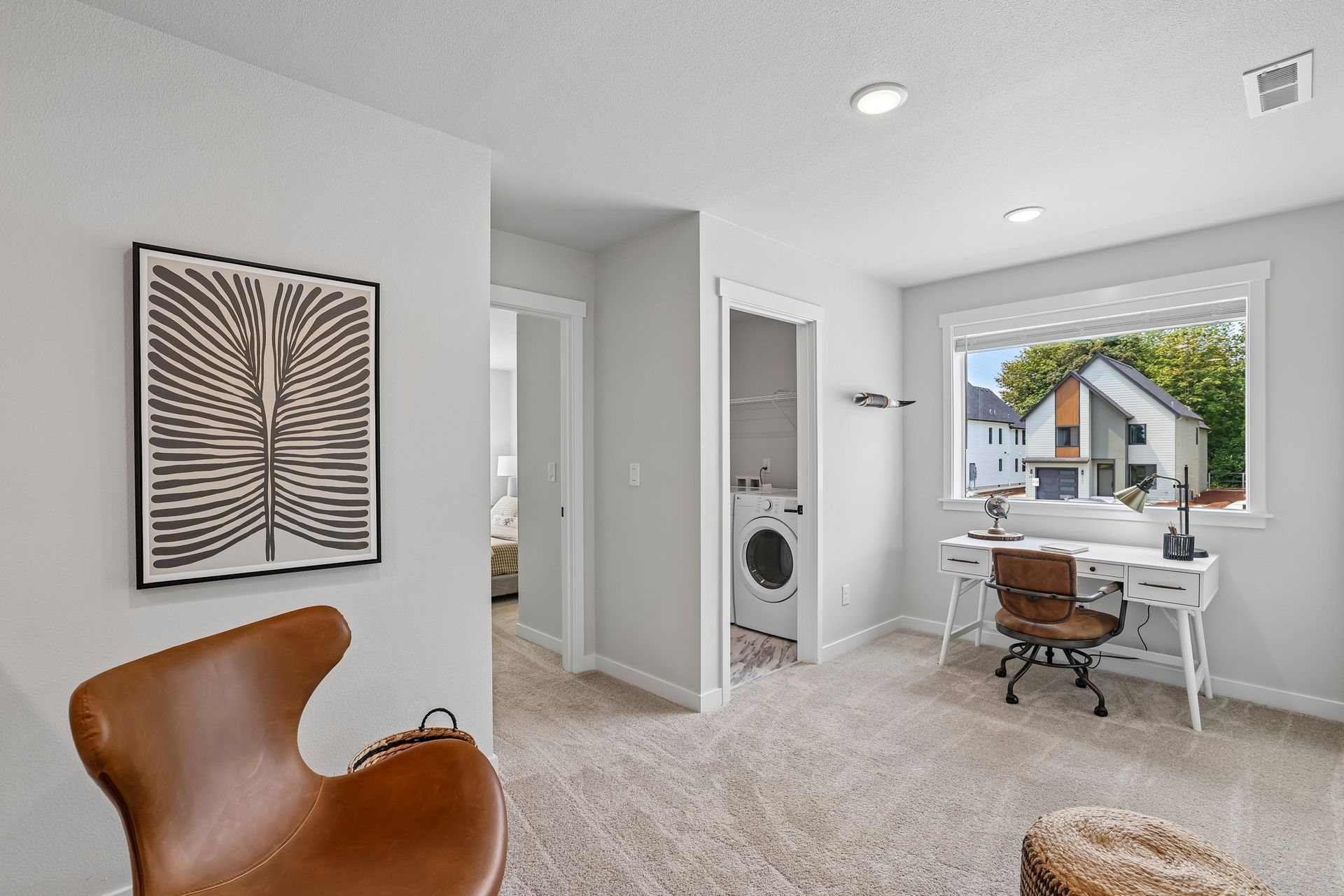 Bright home office with desk, leather chair, laundry nook, and window view of a house outside.