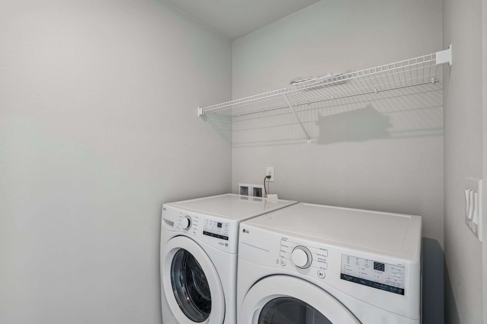 Laundry room with white washer and dryer under a wire shelf in a gray corner.