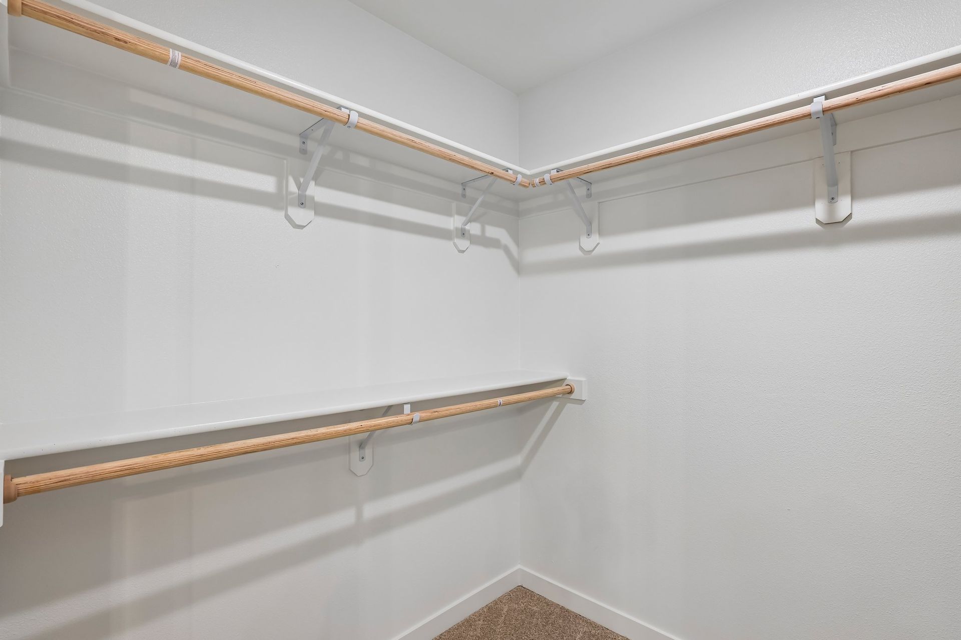 Empty walk-in closet with white walls, beige carpet, and wooden hanging rods on two walls