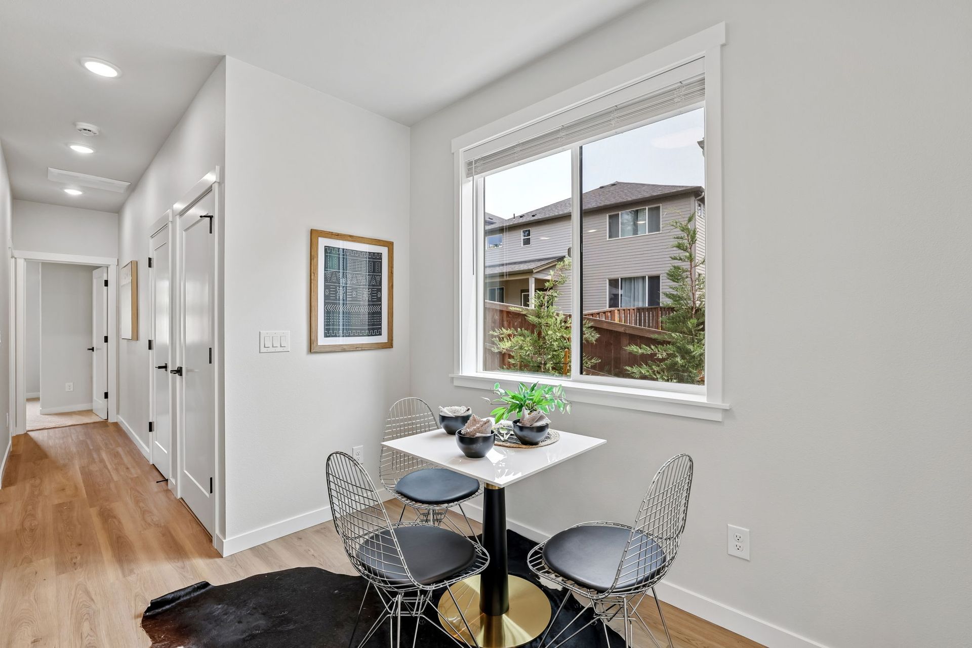 Bright dining nook beside a hallway, with a round table, black chairs, and a window overlooking neighboring houses.