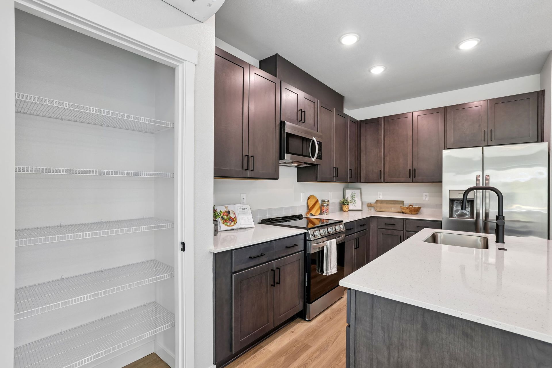 Modern kitchen with dark wood cabinets, white countertops, stainless appliances, and a pantry shelf wall