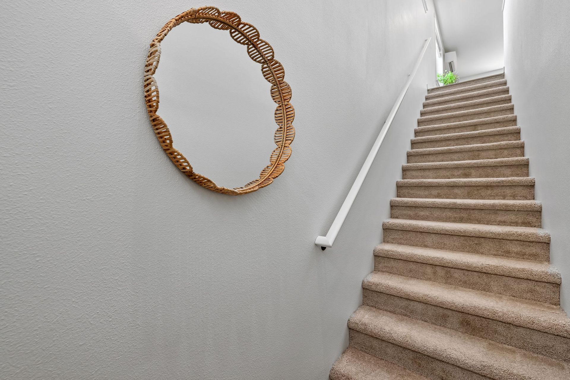 Carpeted stairwell with white walls, a handrail, and a round woven wall mirror.