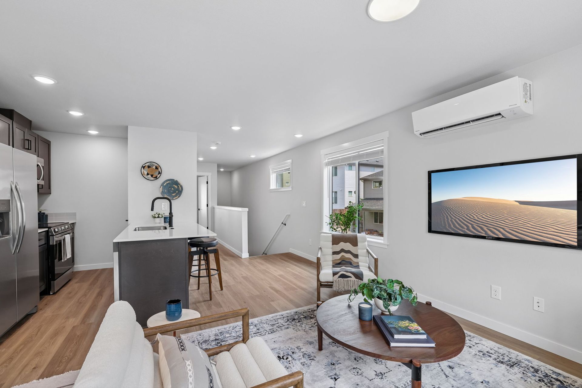 Bright modern living room with kitchen, white walls, wood floors, wall TV, and a plant-filled coffee table.