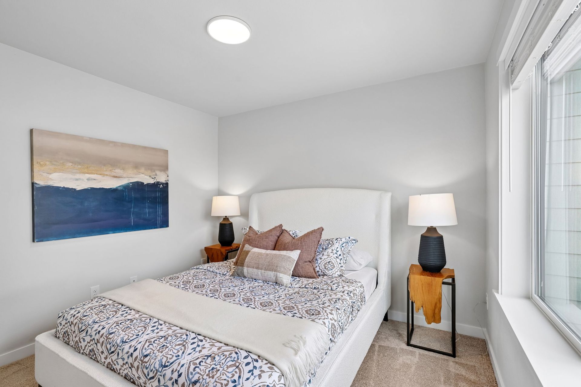 Bright bedroom with a white bed, bedside lamps, abstract wall art, and a large window.