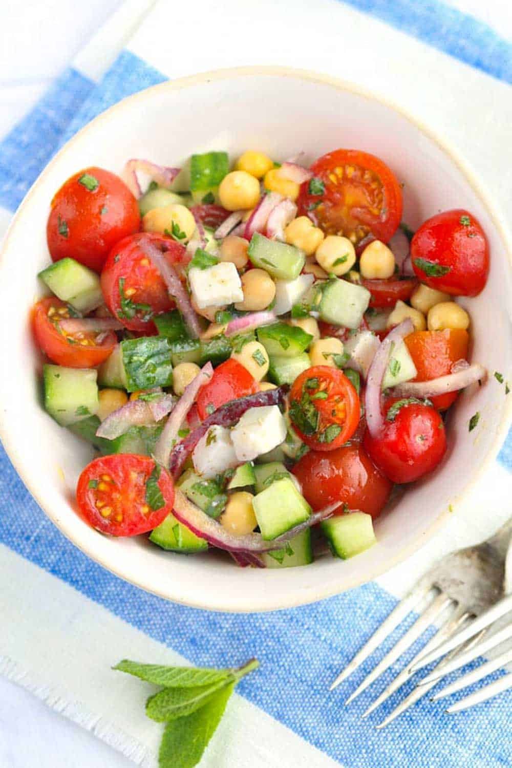 A bowl of salad with tomatoes , cucumbers , chickpeas and onions on a blue and white towel.